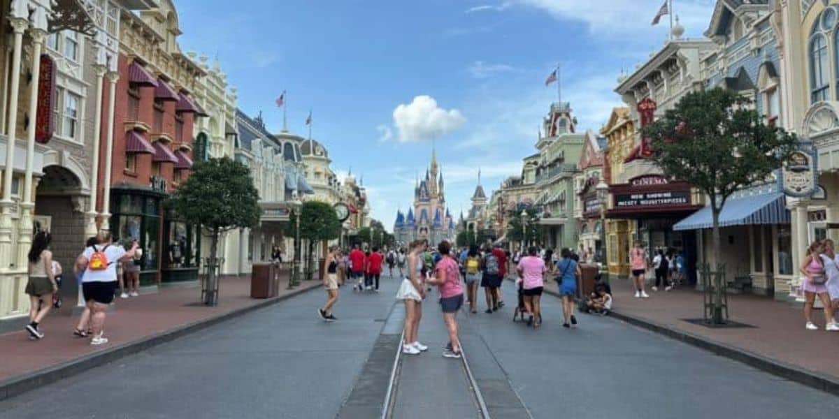 A bustling street scene at Disney World, with a variety of people walking and taking photos. Shops and buildings with decorative facades line both sides. A castle is visible in the distance under a blue sky with scattered clouds.