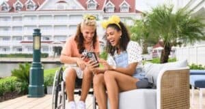 Two women are sitting outside a building with red roofs. One is in a wheelchair, wearing a peach-colored shirt, and the other is on a bench, wearing a white shirt and denim overalls with yellow Minnie Mouse ears. Both are smiling and looking at a smartphone.