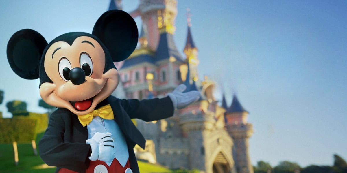 Mickey Mouse, dressed in his classic red shorts, yellow bow tie, and black jacket, stands with one arm outstretched in front of a fairytale castle at Disneyland. The sky is clear and blue, creating a magical, inviting atmosphere.