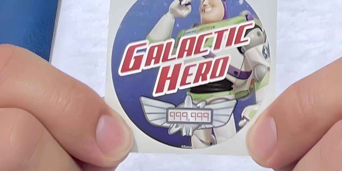 Two hands hold a circular sticker featuring a cartoon astronaut in a white and green spacesuit. The sticker has the text "Galactic Hero" in bold red letters and a score of "999,999" displayed below the astronaut, reminiscent of Buzz Lightyear Space Ranger Spin's high-score achievements.