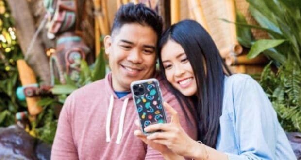 A happy couple takes a selfie together with a smartphone, smiling at the camera. The man wears a red hoodie, and the woman wears a blue shirt. They are outdoors, surrounded by lush greenery and colorful, decorated wooden pillars in the background.