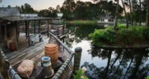 Wide-angle view of Tom Sawyer Island with a rustic wooden dock, barrels, a rowboat, and surrounding greenery along the Rivers of America. Calm water reflects the trees and structures. Various wooden buildings and a boardwalk are visible in the background, creating a tranquil setting.
