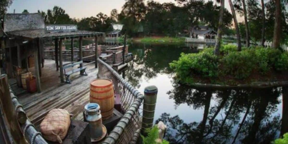 Wide-angle view of Tom Sawyer Island with a rustic wooden dock, barrels, a rowboat, and surrounding greenery along the Rivers of America. Calm water reflects the trees and structures. Various wooden buildings and a boardwalk are visible in the background, creating a tranquil setting.