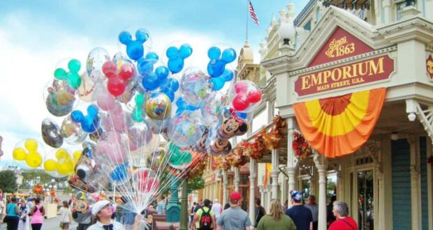A bustling street scene with people walking past the Emporium store on Main Street, USA. A vendor sells colorful balloons, including many shaped like Mickey Mouse heads. The store is decorated with orange and yellow bunting, and an American flag waves in the background.