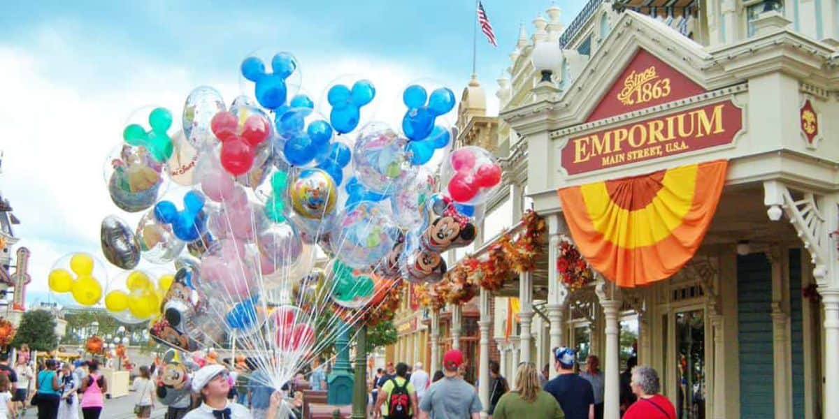 A bustling street scene with people walking past the Emporium store on Main Street, USA. A vendor sells colorful balloons, including many shaped like Mickey Mouse heads. The store is decorated with orange and yellow bunting, and an American flag waves in the background.