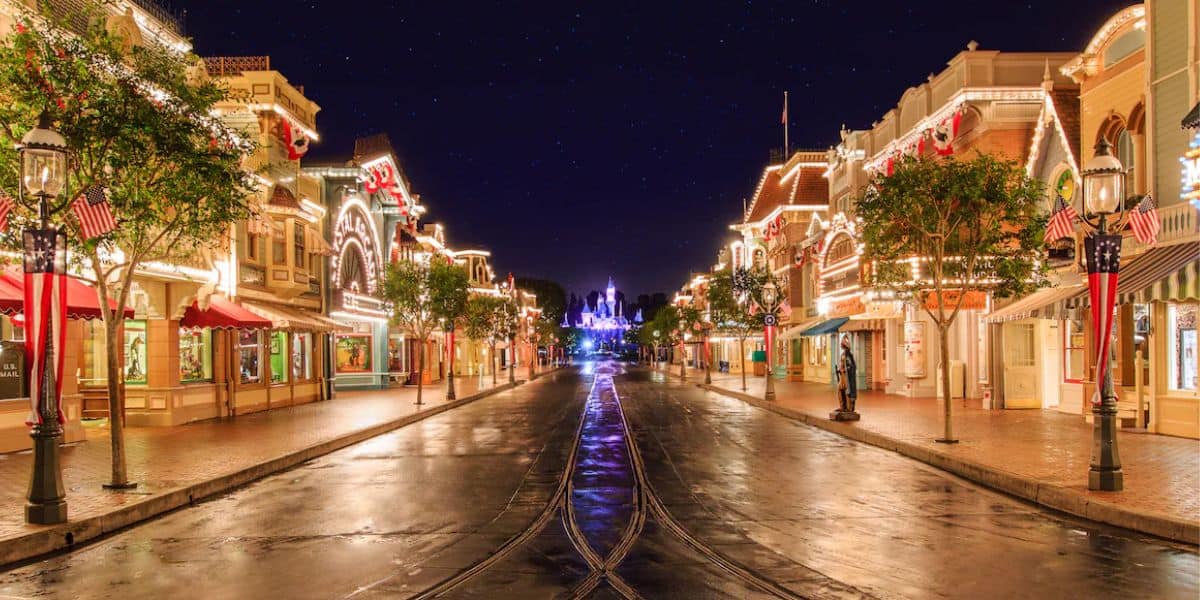A brightly lit, empty Main Street at Disneyland at night. The street is illuminated with festive lights on buildings and lampposts, leading towards Sleeping Beauty Castle in the distance. The wet ground reflects some of the light—perfect for capturing a magical photo or finding an ideal parade viewing spot.
