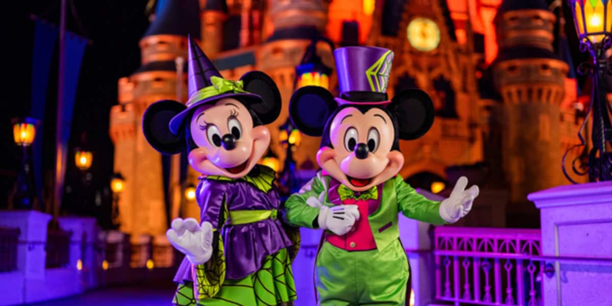 Minnie and Mickey Mouse are dressed in bright green Halloween costumes with a castle illuminated in orange behind them. Minnie wears a witch outfit, and Mickey sports a top hat and bow tie. They stand on a bridge decorated with lights at night, posing for a photo.
