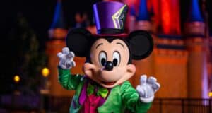 Mickey Mouse dressed in a colorful Halloween outfit, including a green jacket, pink vest, and purple top hat, poses with outstretched hands in front of a brightly lit castle at night. The background features vibrant red and blue lighting on the castle.