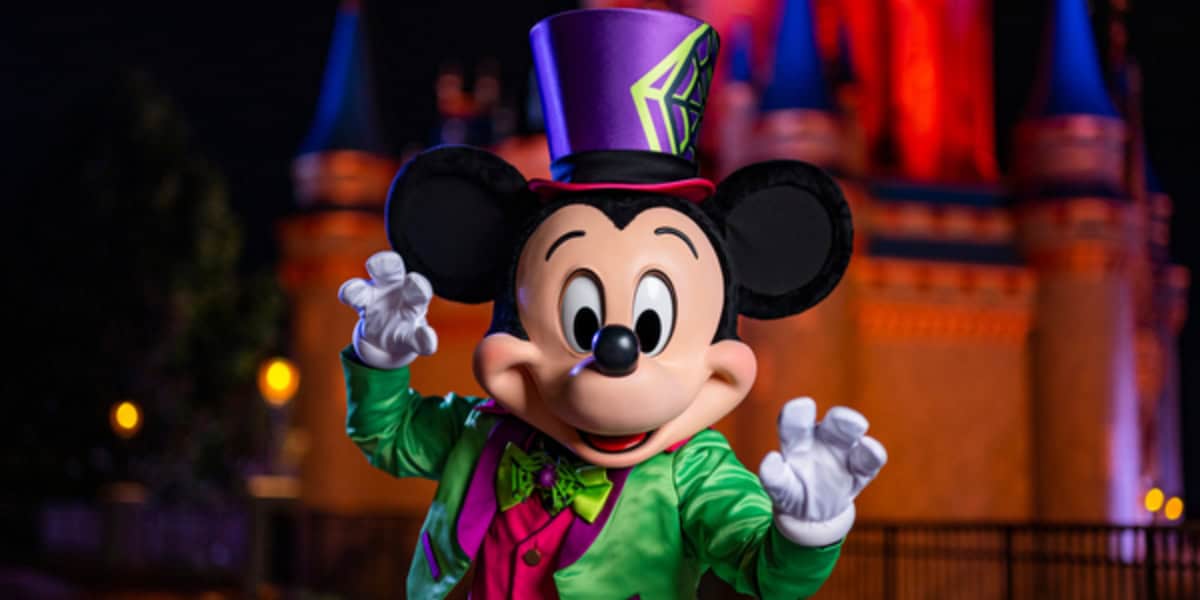Mickey Mouse dressed in a colorful Halloween outfit, including a green jacket, pink vest, and purple top hat, poses with outstretched hands in front of a brightly lit castle at night. The background features vibrant red and blue lighting on the castle.