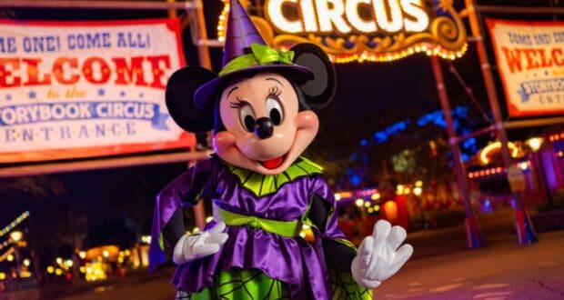 Minnie Mouse is dressed in a vibrant purple and green witch costume with an orange bow, smiling and posing in front of a brightly lit circus-themed background with banners reading "Welcome to the Storybook Circus." The scene is colorful and festive.