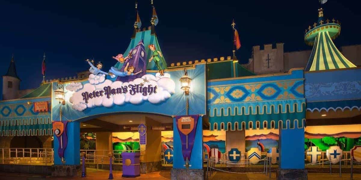 Photo of the entrance to the Peter Pan's Flight ride at a theme park. The entrance, adorned with colorful decorations featuring Peter Pan, Wendy, John, and Michael flying, is illuminated by lanterns against the dark night sky.