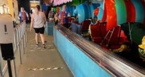 A few people stand in line at an indoor amusement park ride inspired by Peter Pan. The ride vehicles are colorful and whimsical with an overhead canopy. Social distancing markers are on the ground, and a hand sanitizer station is visible on the left.