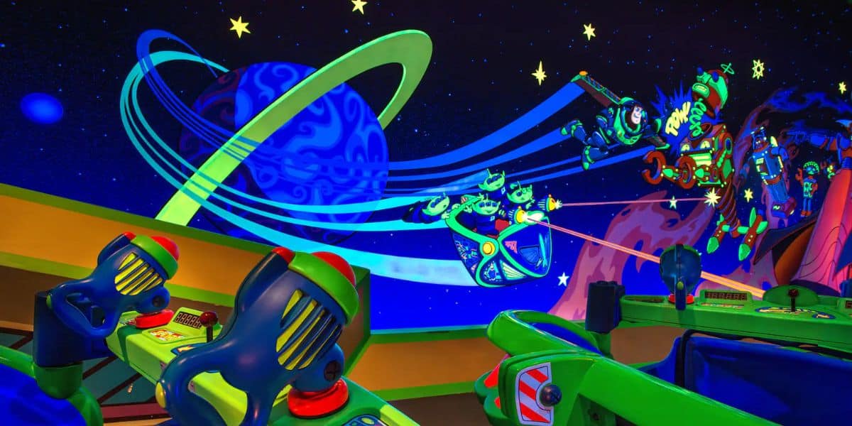 Image of a vibrant, space-themed arcade attraction with neon colors. Toy ray guns and control panels are visible in the foreground, while the background features cosmic scenery with planets, stars, and animated characters engaged in a space battle reminiscent of Buzz Lightyear Space Ranger Spin.