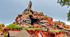 A colorful and detailed rock formation resembling a mountain, with rugged terrain, patches of greenery, and a distinct peak. The sky is overcast, adding contrast to the vibrant hues of the rocks. At its base, a wooden structure with a peaked roof hints at the Magic Kingdom's newest attraction launching next month.