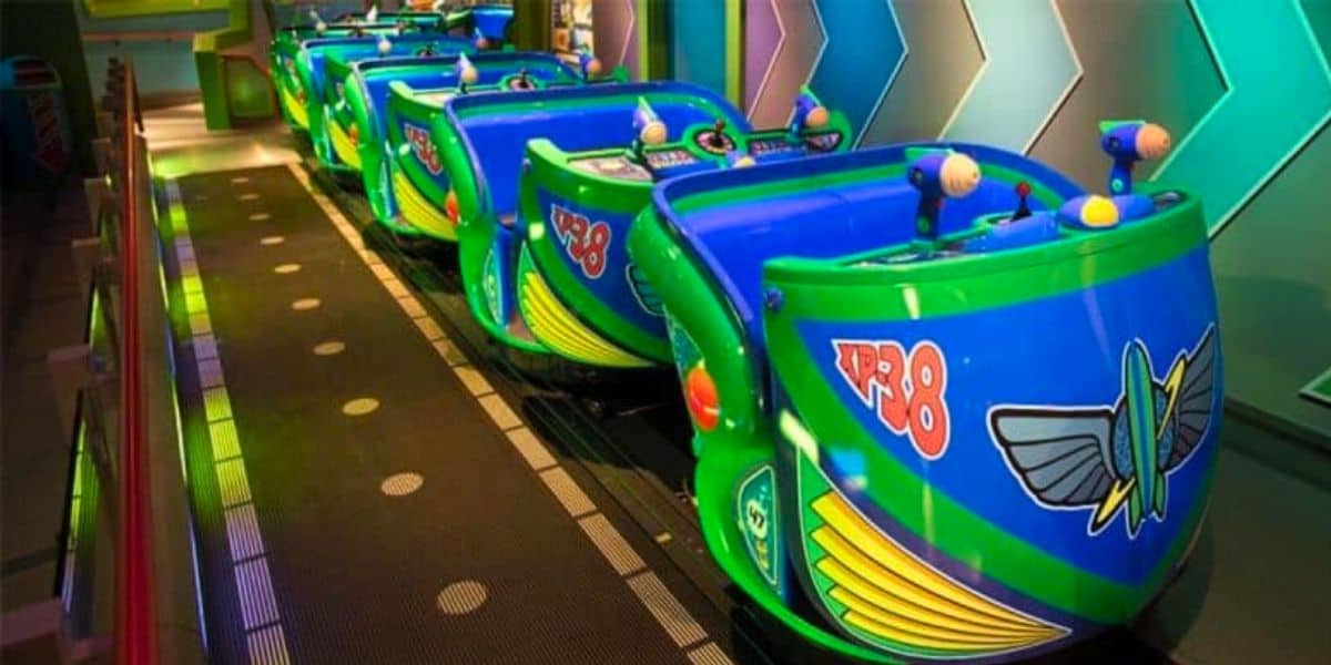 Four turquoise and green ride vehicles, themed after Toy Story spacecraft and featuring "XP-38" decals, are lined up on a track, ready for passengers. They have seats and twin laser blasters, set against a futuristic backdrop with zigzag neon patterns.