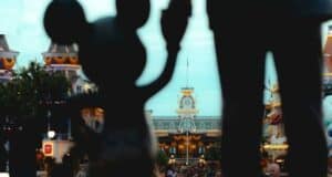 Silhouette of Mickey Mouse holding hands with a statue, set against a backdrop of a Disney theme park entrance with decorative buildings and a busy crowd below. Bright, festive lighting enhances the cheerful atmosphere while celebrating unity and diversity.