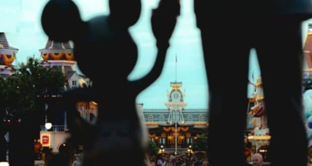 Silhouette of Mickey Mouse holding hands with a statue, set against a backdrop of a Disney theme park entrance with decorative buildings and a busy crowd below. Bright, festive lighting enhances the cheerful atmosphere while celebrating unity and diversity.
