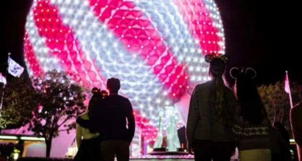 A nighttime scene at Epcot with a family of four wearing Mickey Mouse ears admiring the large, illuminated Spaceship Earth. The geodesic sphere is lit up with bright, colorful patterns, primarily in red and white, creating a festive atmosphere.