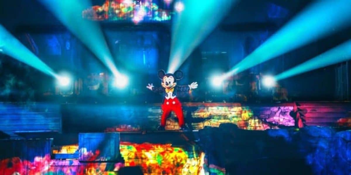Mickey Mouse stands center stage with arms outstretched, illuminated by vibrant, colorful lights and surrounded by smoke and bright beams in a theatrical setting. The background features diverse, moving patterns of light and colors, creating a magical atmosphere.
