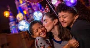 A joyful mother kisses one of her two smiling children, both wearing Mickey Mouse ear headbands, in front of a colorful, illuminated castle at night during Disney After Hours.