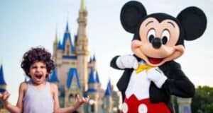 A young child with curly hair, wearing a sleeveless shirt, stands excitedly next to a person in a Mickey Mouse costume. Behind them, there is a large castle with blue and gold accents, reminiscent of a fairy tale setting. Both appear to be at a theme park.