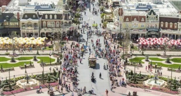 A bustling theme park with visitors walking along a main street lined with colorful buildings and decorative awnings. On both sides of the street are neatly arranged gardens, fountains, and seating areas. The scene is lively and vibrant under a clear sky.