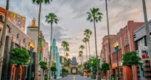 A picturesque street lined with tall palm trees and charming buildings on either side. The buildings have a vintage, Hollywood-inspired architectural style. The sky is mostly cloudy with hints of sunlight. The street appears peaceful and empty.