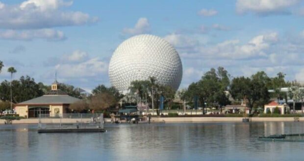 A large white geodesic sphere, known as Spaceship Earth, stands prominently in the background at Epcot theme park in Disney World. The foreground features a calm body of water reflecting the sphere, with buildings and trees on the shoreline under a partly cloudy sky.