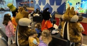Several children are sitting on black benches, watching a cartoon on a screen in a room with star decorations on the wall. Adults are present in the background. Three people in large, plush animal costumes are seated with the children, two resembling chipmunks.