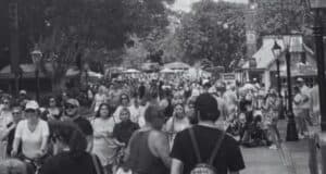 A bustling crowd of people walks along a tree-lined outdoor path, possibly in an amusement park or public space. The scene is in black and white, with various individuals engaging in different activities. Small structures and lamp posts line the path.
