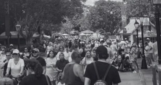 A bustling crowd of people walks along a tree-lined outdoor path, possibly in an amusement park or public space. The scene is in black and white, with various individuals engaging in different activities. Small structures and lamp posts line the path.
