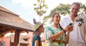 A family enjoys their time outdoors. A young child wearing a hat with mouse ears looks up at a woman holding a smartphone, who is smiling, perhaps using the chat feature on Walt Disney World's website. A man in a white shirt stands next to them, also smiling. They are near a thatched-roof building and tall palm trees.