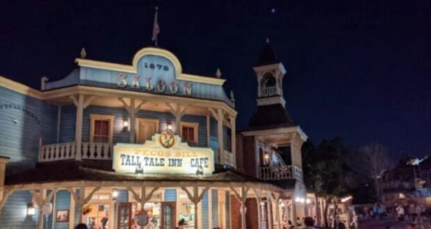 A nighttime photo of a western-themed building with a sign that reads "Pecos Bill Tall Tale Inn and Cafe" and "Saloon." The building has a rustic facade and an old-fashioned clock tower. People are walking around the area. The sky is dark with a few visible stars.