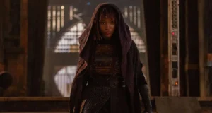 A person dressed in dark clothing and a hooded cape stands in a dimly lit, industrial setting. The individual is looking forward with a serious expression. The background includes metallic structures and arched openings illuminated by soft light.