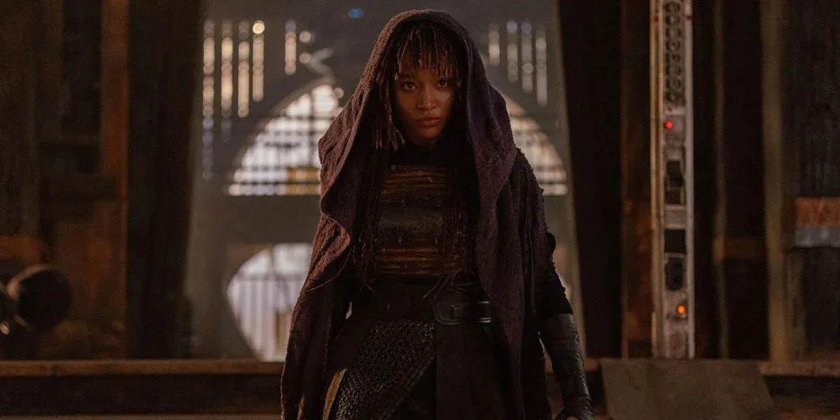 A person dressed in dark clothing and a hooded cape stands in a dimly lit, industrial setting. The individual is looking forward with a serious expression. The background includes metallic structures and arched openings illuminated by soft light.