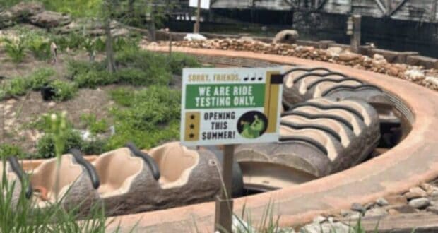 A water ride with log-shaped boats is under testing, indicated by a sign that reads, "Sorry, friends. We are ride testing only. Opening this summer!" The ride's serpentine track is visible, surrounded by greenery. Some structures and a bridge are in the background.