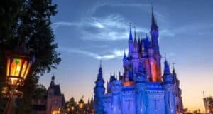 The image shows a majestic, illuminated castle at dusk, likely from a famous theme park, with its towers and spires glowing against a deep blue sky. A vintage lamppost with multicolored glass panels is visible on the left, adding to the enchanting atmosphere.