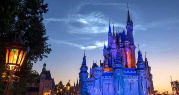 The image shows a majestic, illuminated castle at dusk, likely from a famous theme park, with its towers and spires glowing against a deep blue sky. A vintage lamppost with multicolored glass panels is visible on the left, adding to the enchanting atmosphere.
