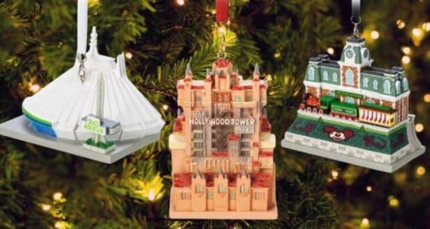 Three theme park ride ornaments hang on a Christmas tree. They depict a futuristic white structure, The Hollywood Tower Hotel, and a train station with a green train. The tree is decorated with warm white lights.