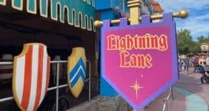A sign for "Lightning Lane" is displayed against a colorful medieval-themed backdrop, featuring red and white striped and blue shield decorations. People walk in the background under a partly cloudy sky.