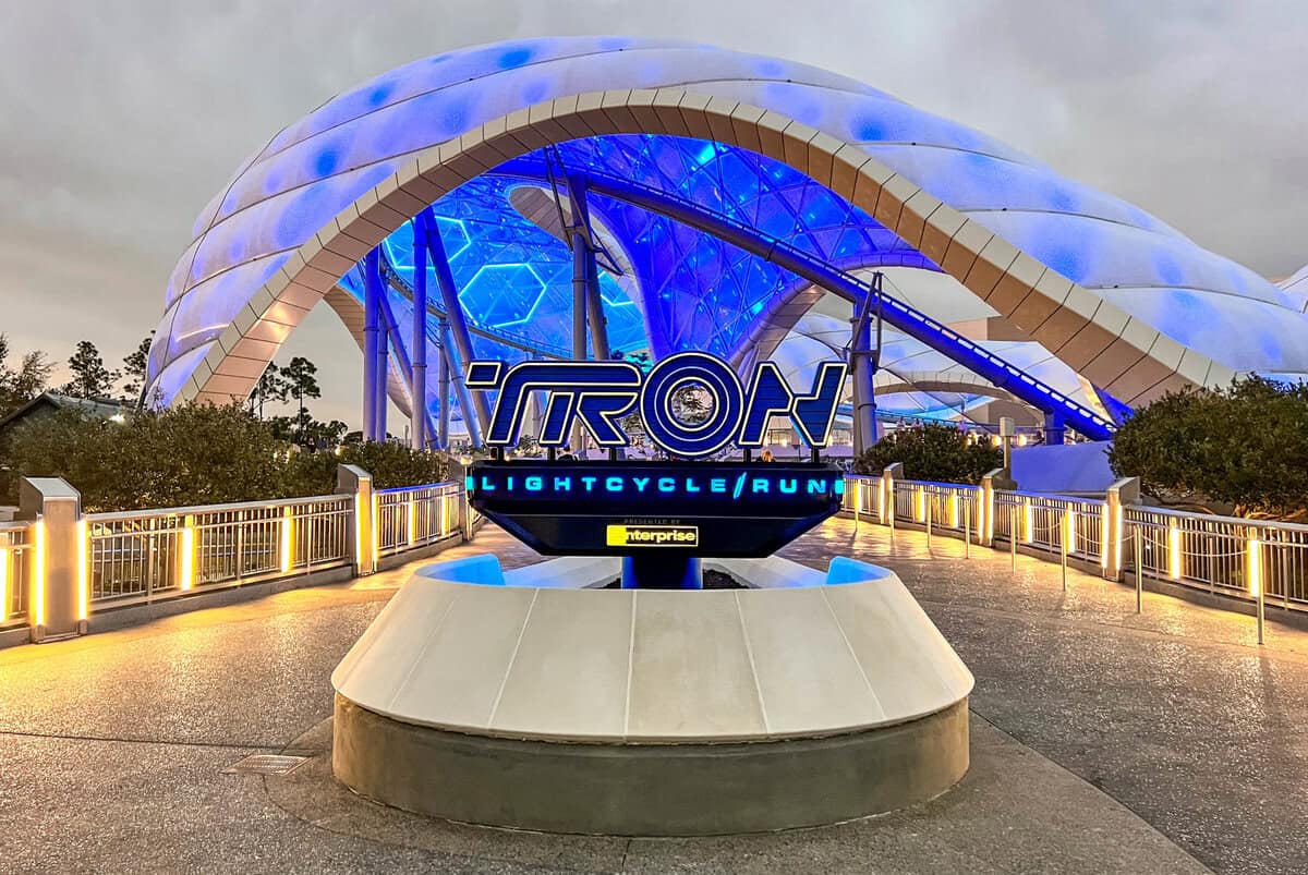The image shows the entrance to the TRON Lightcycle Run attraction at Magic Kingdom, featuring a futuristic structure with blue and white lighting. A curved canopy covers the area, providing shade near a convenient water refill station, while a prominent sign displays the attraction's name.