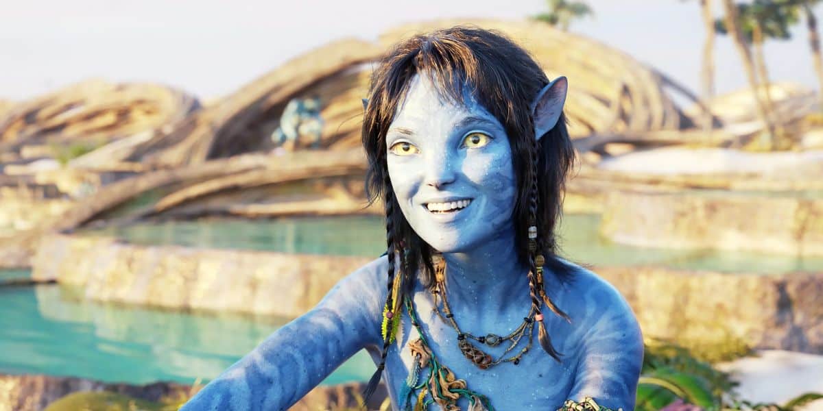 A blue, humanoid character with pointed ears and yellow eyes smiles amidst water and twisted tree-like structures in Avatar: The Way of Water.
