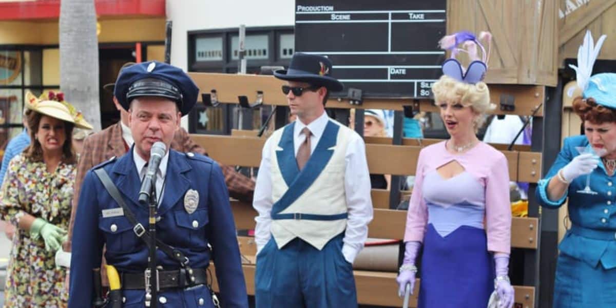 A group of people dressed in vintage 1940s attire are gathered outside, capturing the essence of Hollywood Studios. A man in a police uniform stands at the front holding a microphone, while others in stylish costumes and hats look on. A clapperboard is visible in the background.