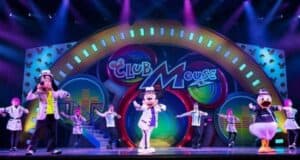 Colorful stage performance with cartoon characters, including a dog, a mouse, and a duck, wearing lab coats and dancing with human performers. Bright lights and vibrant "Club Mouse" sign in the background.