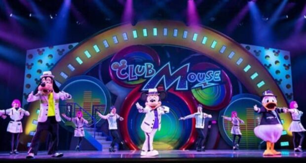 Colorful stage performance with cartoon characters, including a dog, a mouse, and a duck, wearing lab coats and dancing with human performers. Bright lights and vibrant "Club Mouse" sign in the background.