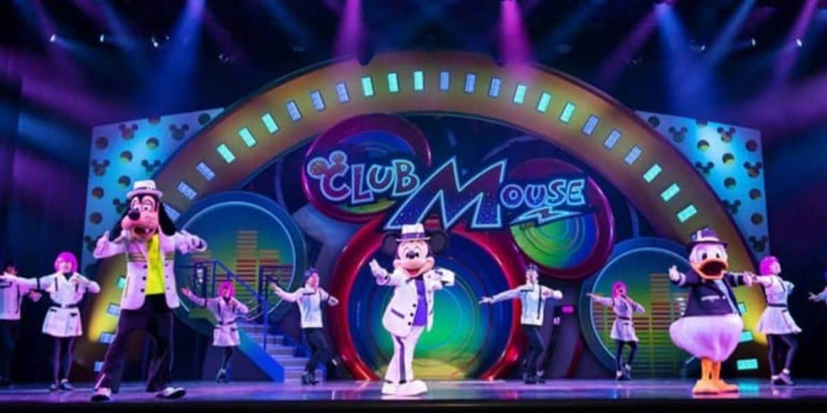 Colorful stage performance with cartoon characters, including a dog, a mouse, and a duck, wearing lab coats and dancing with human performers. Bright lights and vibrant "Club Mouse" sign in the background.