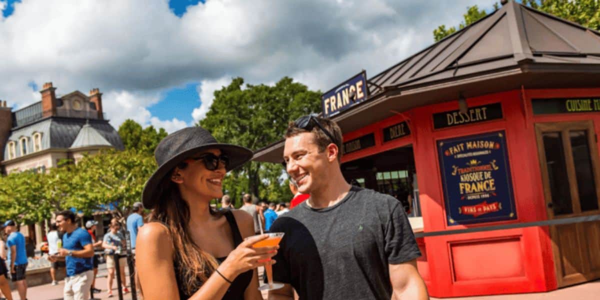 A couple smiles and holds drinks in front of a red food booth labeled "France" at an outdoor festival. The scene is lively with people walking around, and there are trees and a building in the background under a partly cloudy sky.