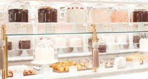 A display case filled with various elegant cakes and pastries, each covered by glass domes. The cakes are arranged on white stands, featuring different flavors and frosting styles, in a bright and inviting bakery setting.