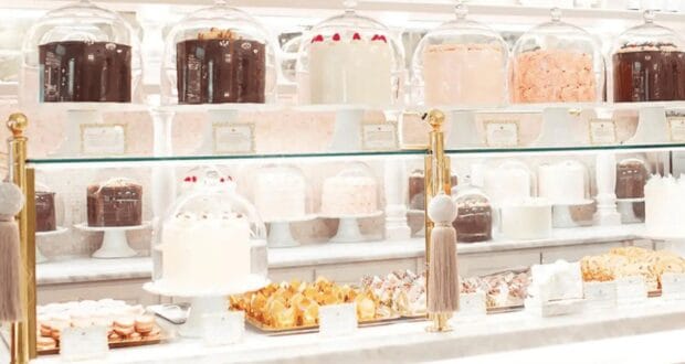 A display case filled with various elegant cakes and pastries, each covered by glass domes. The cakes are arranged on white stands, featuring different flavors and frosting styles, in a bright and inviting bakery setting.