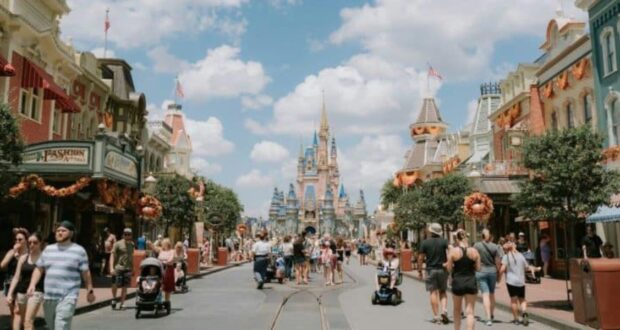 People stroll along a lively street lined with shops adorned with autumn decorations. In the background, a fairy tale castle stands under a blue sky with fluffy clouds. Families and tourists enjoy the bustling atmosphere.